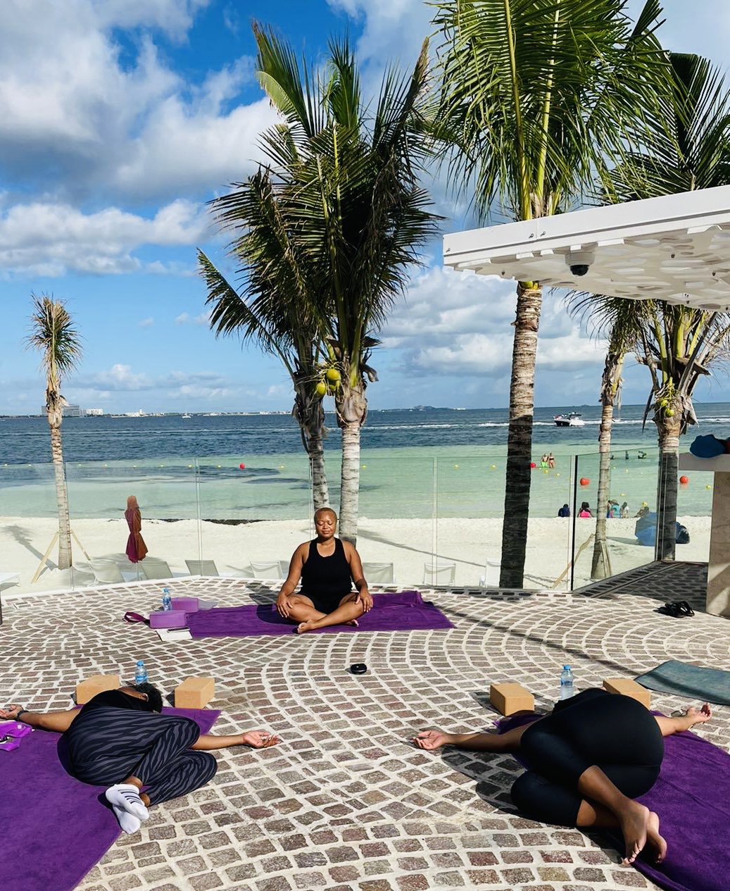 Instructor leading a beach yoga class with participants on purple mats, turquoise water and palm trees in background